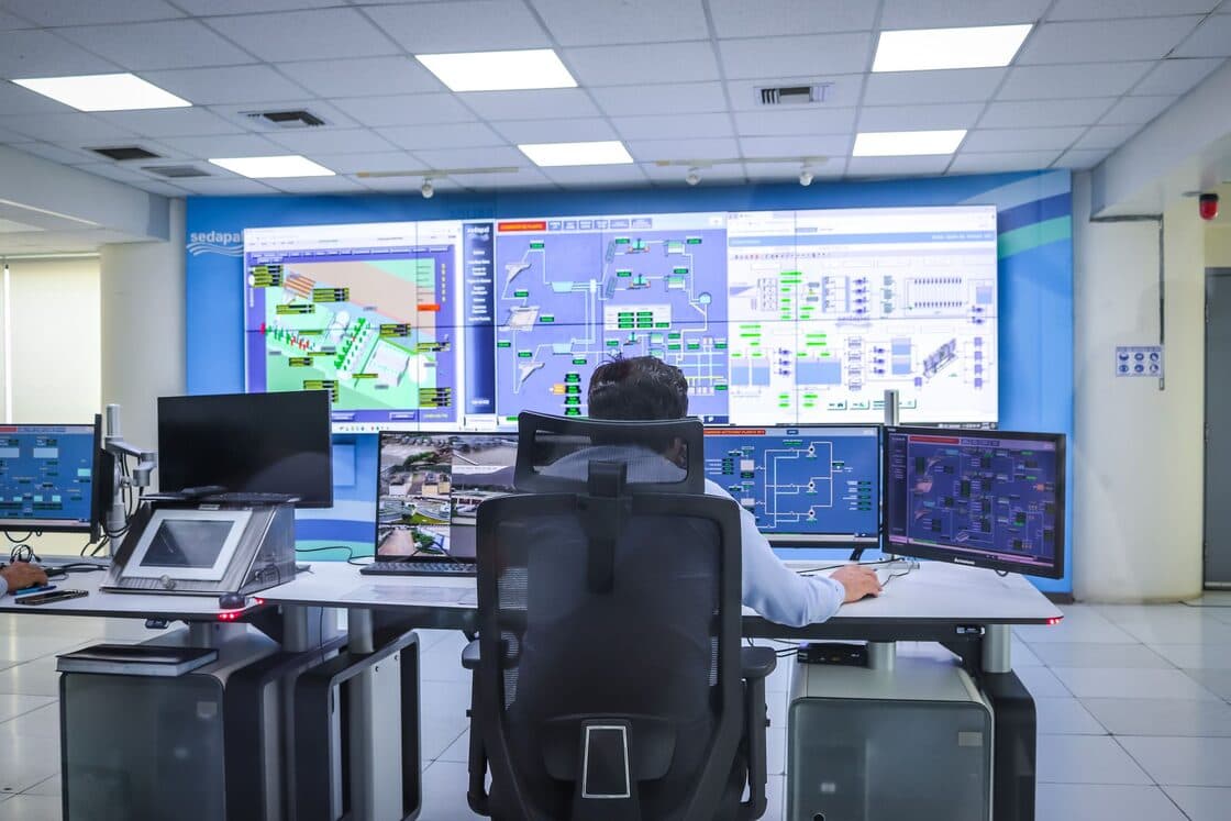 Operator at a control-room desk facing a wall of mimic diagrams and monitoring screens