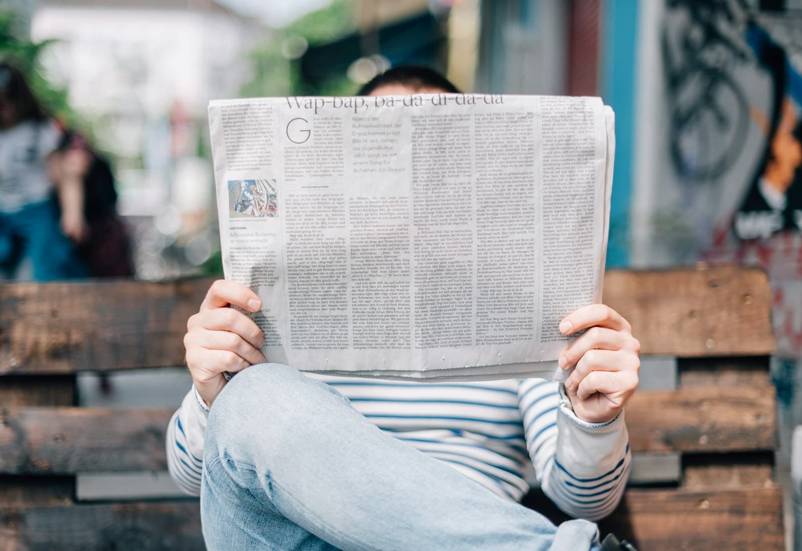 Person seated on a wooden bench reading a printed newspaper.